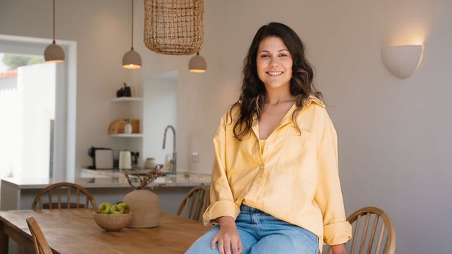 A smiling young woman in a yellow shirt and blue jeans sitting on a wooden dining table in a modern, open-concept kitchen and dining room.