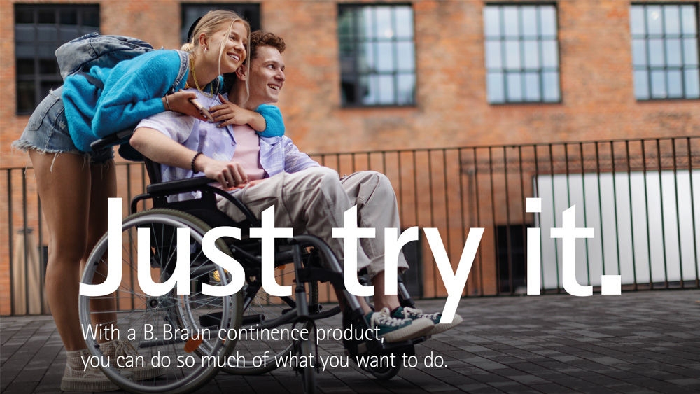 A teenage girl in a blue jacket and shorts is hugging a teenage boy sitting in a wheelchair outdoors. They are on a paved area with a brick building in the background. The text on the image reads: "Just try it. With a B. Braun continence product, you can do so much of what you want to."