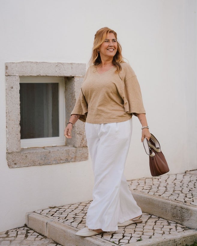 A smiling woman with blonde hair, wearing a beige v-neck top and wide-leg white trousers, walks down a stone-paved sidewalk past a white wall with a stone-framed window while carrying a brown leather handbag.