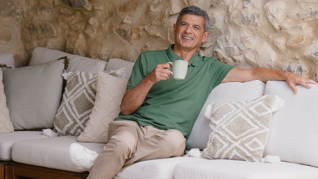 A smiling man sits comfortably on an outdoor sofa with white cushions, holding a ceramic mug. He is wearing a green polo shirt and beige trousers, leaning back against a stone wall background.