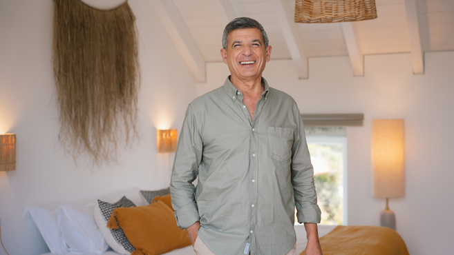 A smiling confident man wearing a green shirt, standing in a modern, brightly lit bedroom featuring warm lighting, a woven wall hanging, and yellow mustard colored bedding.