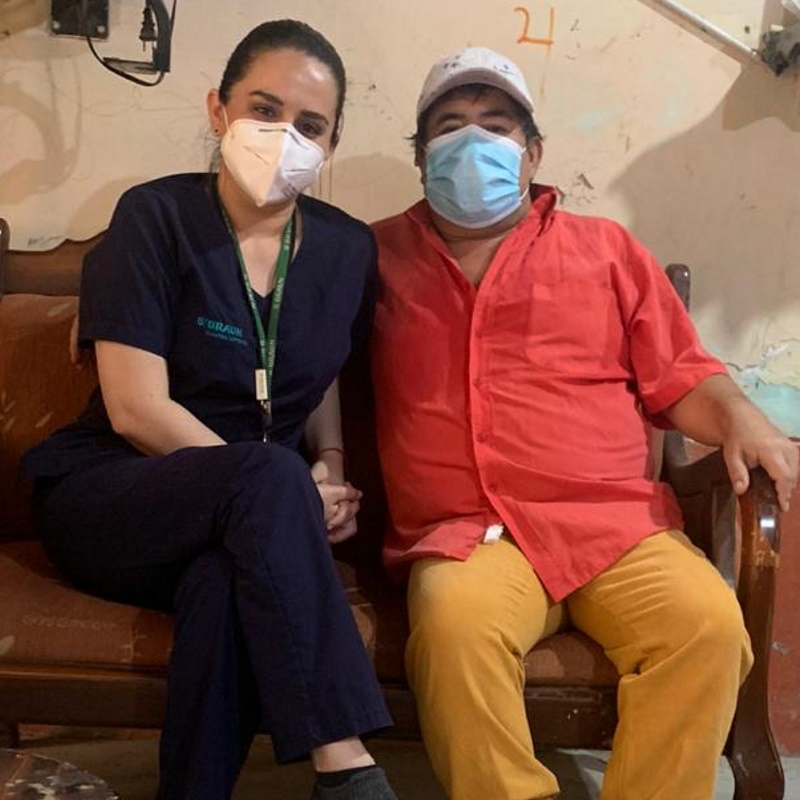 Carol sits next to Carlo on a sofa, both looking into the camera and wearing face masks.