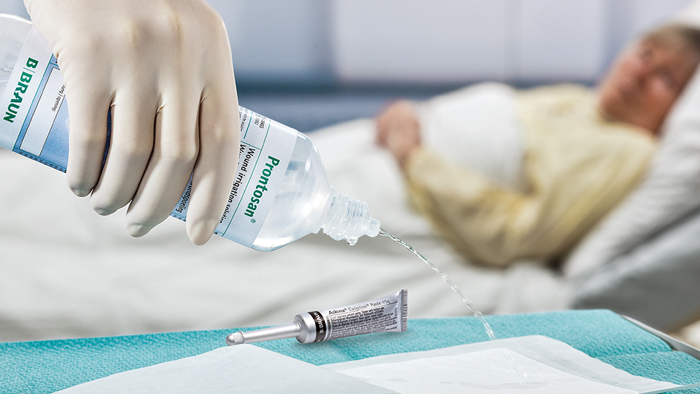 A hand with a glove holds a bottle of prontosan and squeezes the liquid onto a large plaster. An askina tube lies on the table. A patient lies in bed in the background.