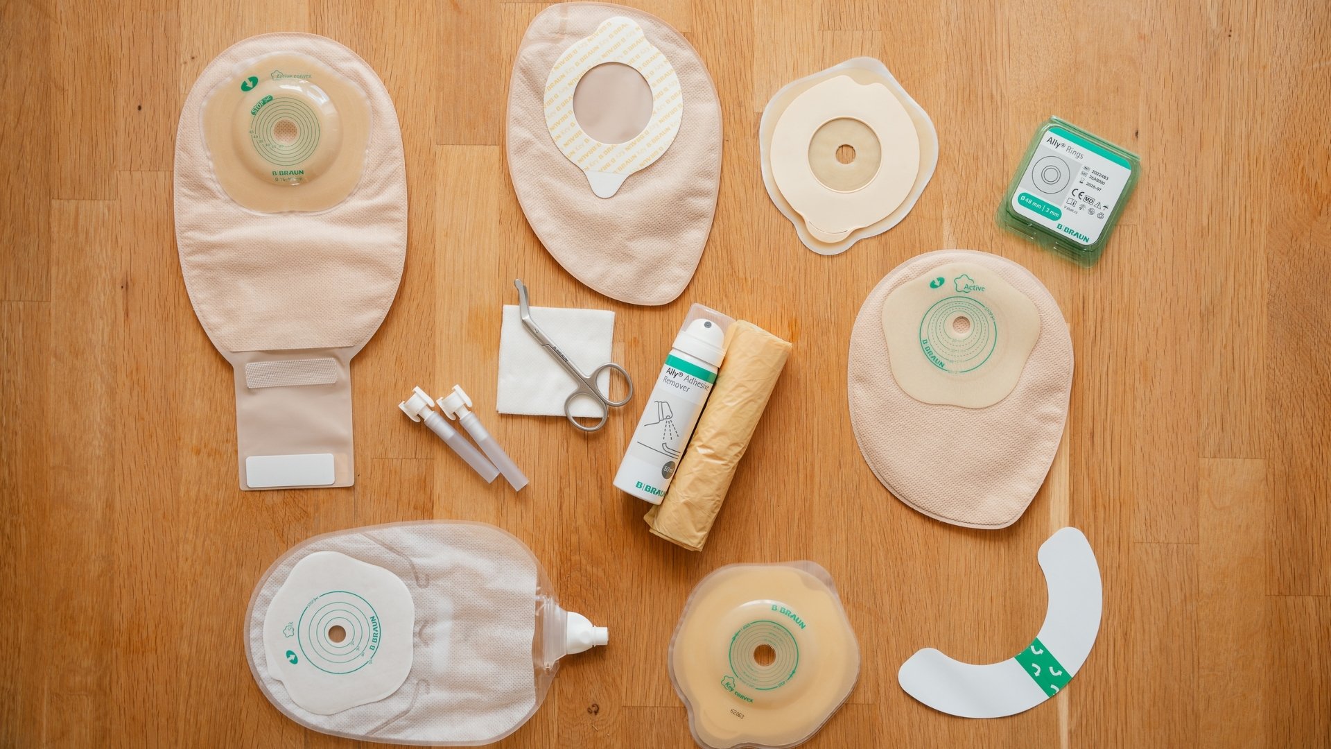 A flat lay overview of B. Braun stoma products and accessories arranged on a light wooden surface. The collection includes different types of stoma beige bags, stoma wafers, skin barriers, stoma rings, curved medical scissors, adhesive remover spray, and disposal bag.