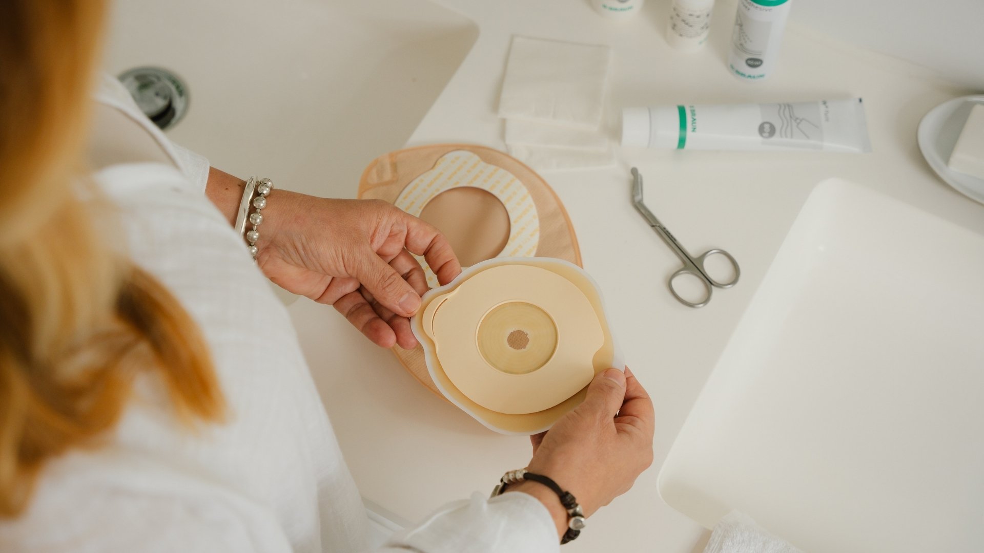An overhead view of a person holding a stoma wafer over a white bathroom counter. In the background, a beige stoma bag, medical scissors, and stoma accessories.